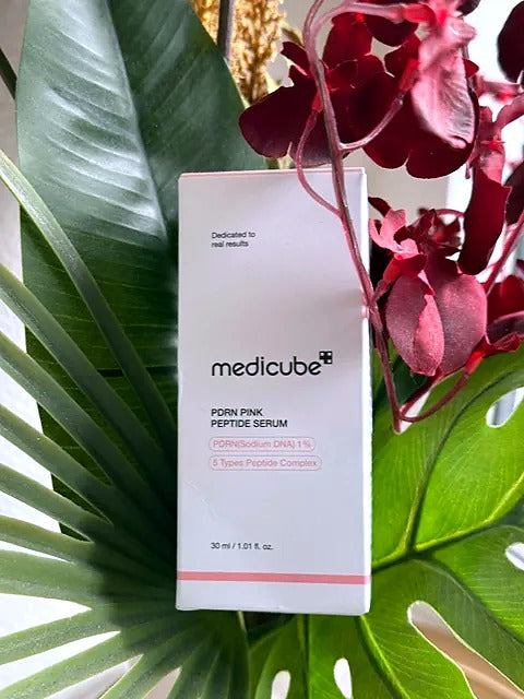 Medicube PDRN Pink Peptide Serum i æske mellem grønne blade og røde blomster